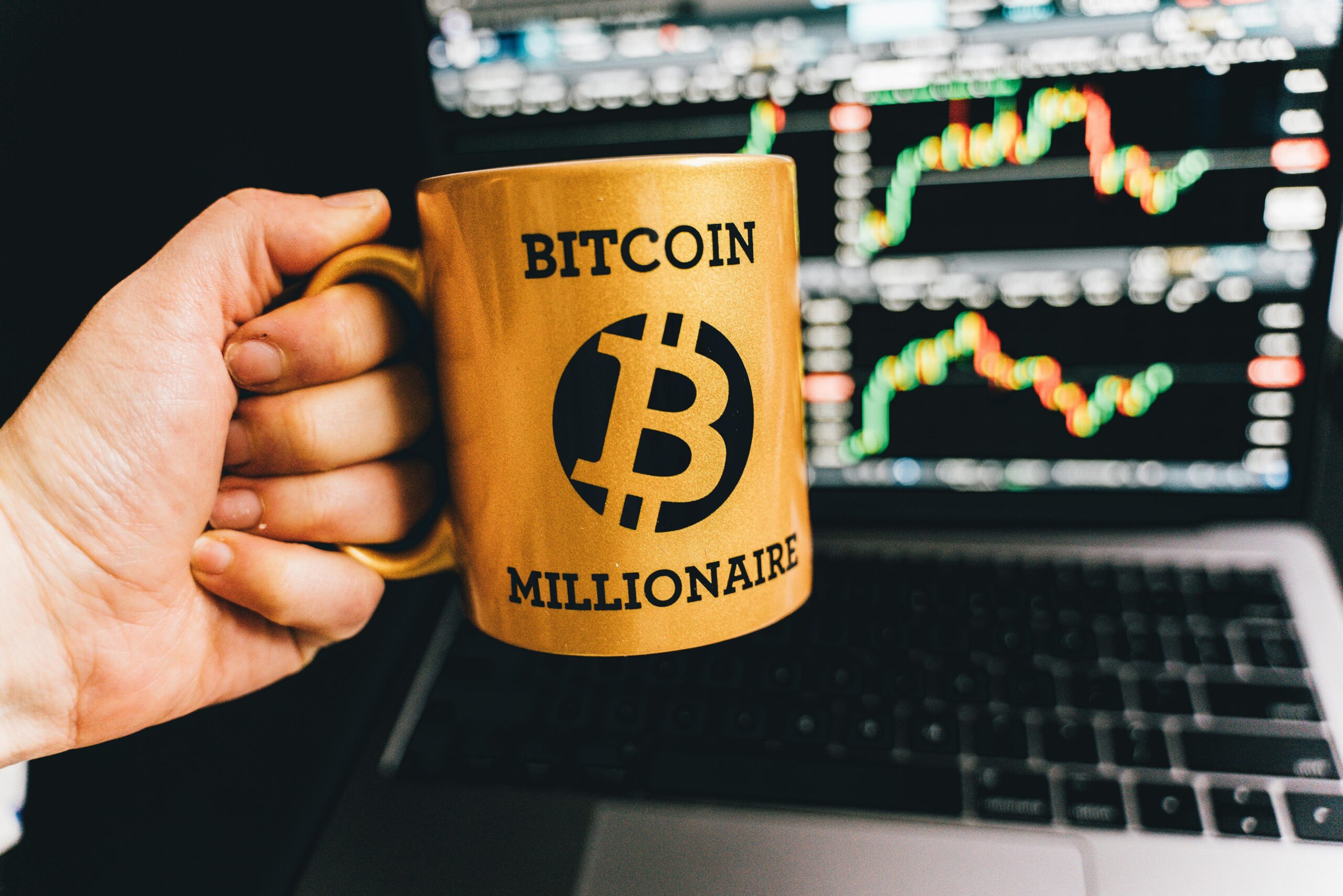 Golden mug with 'Bitcoin Millionaire' text in front of stock market charts on laptop screen.